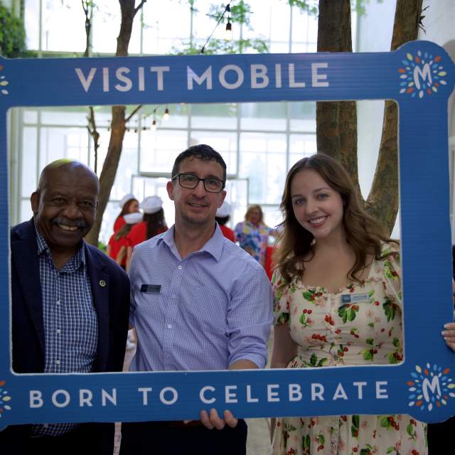 two men and a woman in a hand-held selfie frame that reads Visit Mobile, Born to Celebrate