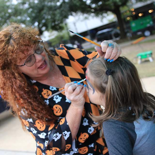 Woman dressed in halloween-themed clothes painting the face of a child.
