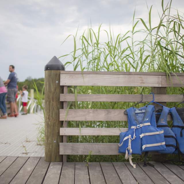 people walking down the boardwalk to the beach, fence with life jackets hanging