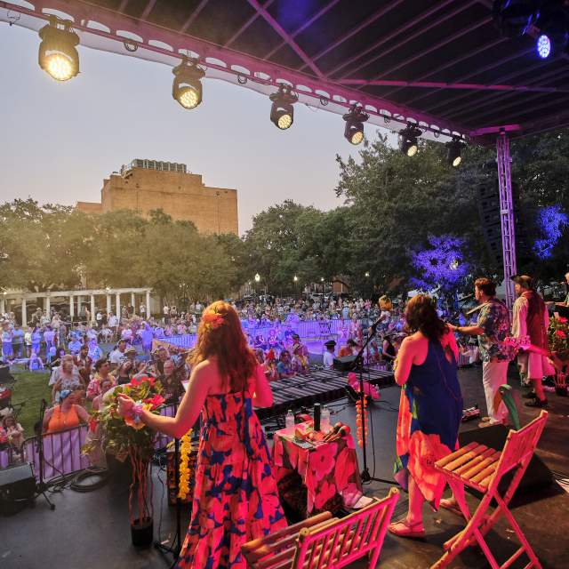 Group of singers and musicians dressed in tropical, summer attire performing in front of a crowd on stage