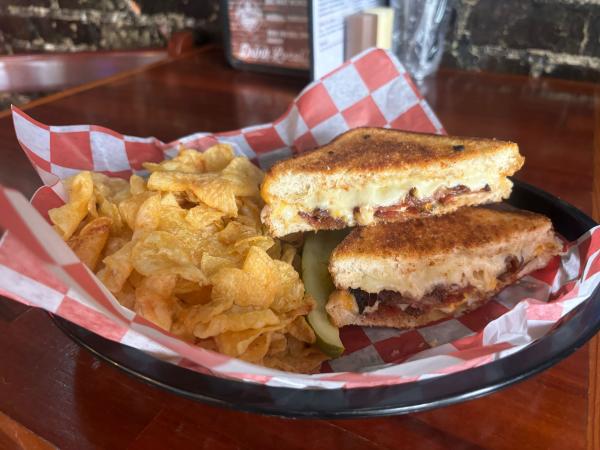 Grilled cheese with bacon, cut in half with chips and a pickle in a basket with red and white checkerboard paper