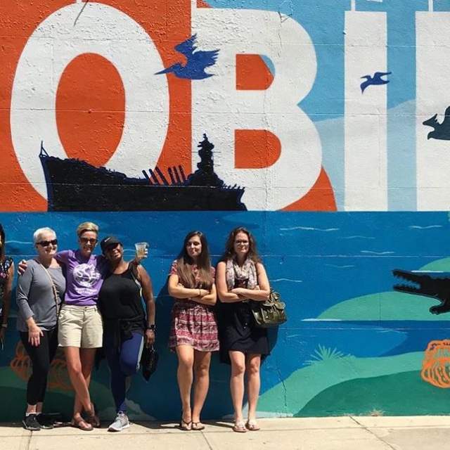 Group of women on the sidewalk in front of the Mobile mural of the USS Alabama and an alligator