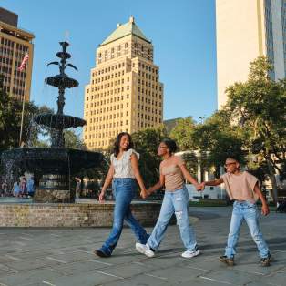 Two Black girls and one Black boy walk in a park past a fountain with buildings in the background