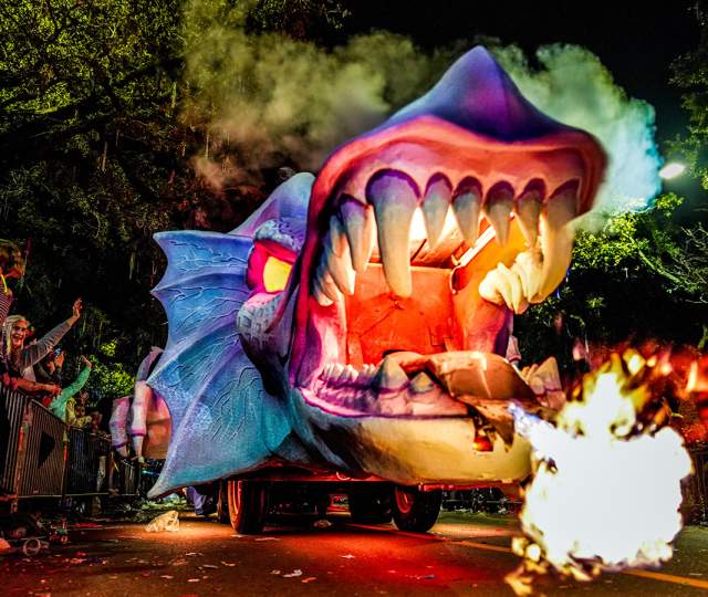 A purple and blue Mardi Gras dragon float breathes fire while on a parade route