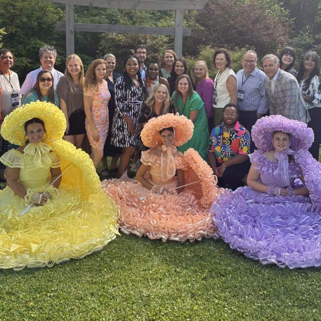 A group of business men and women stand in a line behind girls in pastel dresses seated