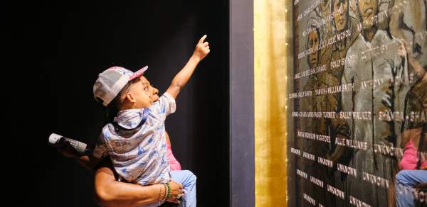 African American man holding a young child in front of the plaque with engraved names of the Clotilda, the Last American Slave Ship