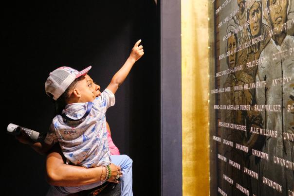 African American man holding a young child in front of the plaque with engraved names of the Clotilda, the Last American Slave Ship